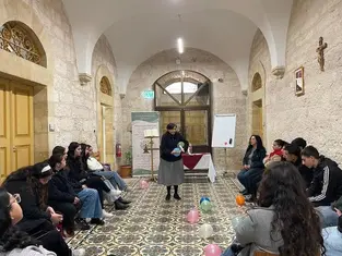 A nun speaks to seated people in a stone room with colorful balloons on patterned floor. An exit sign, posters, and a cross adorn the walls.
