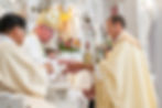 Two clergymen in ceremonial robes perform a ritual. Ornate church interior with flowers in the background. Solemn mood.