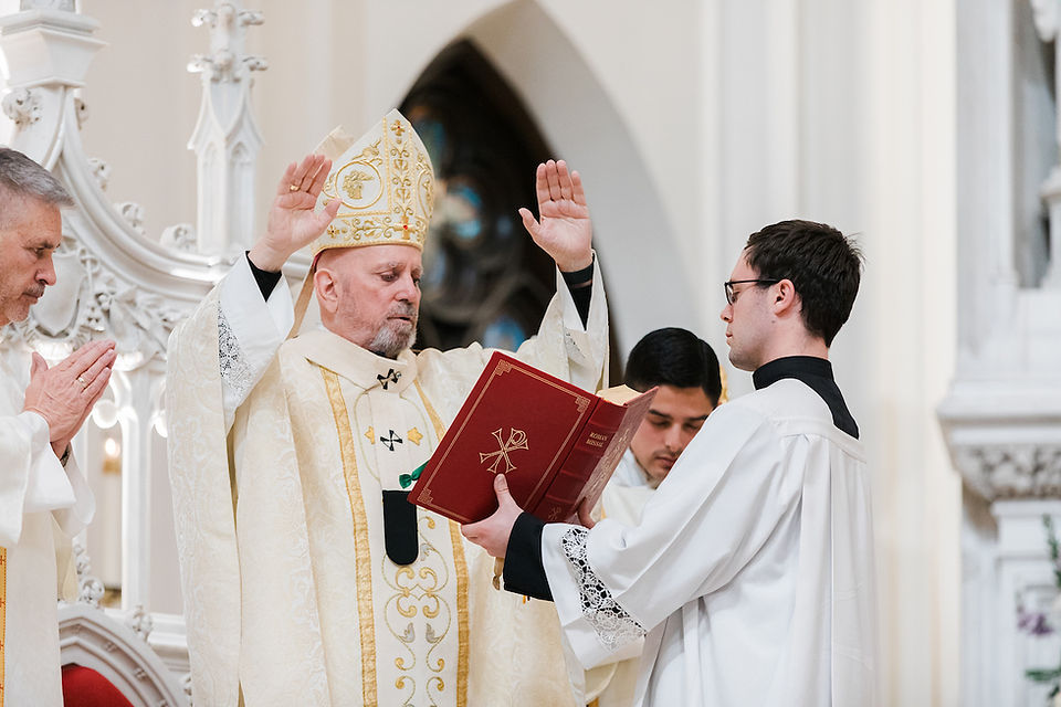 A bishop in gold vestments raises hands in blessing, a priest holds a red book labeled Roman Missal. Ornate church setting.