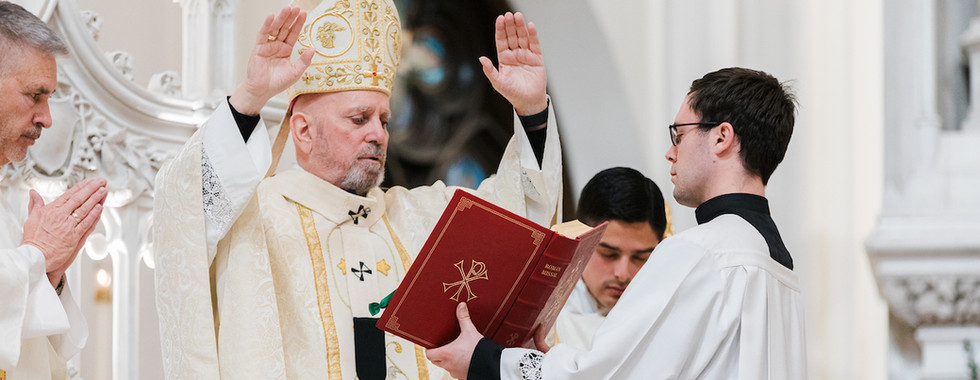 A bishop in gold vestments raises hands in blessing, a priest holds a red book labeled Roman Missal. Ornate church setting.
