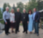 Group of six people smiling on a stone patio, wearing formal and casual attire, with lush green trees and overcast sky in the background.