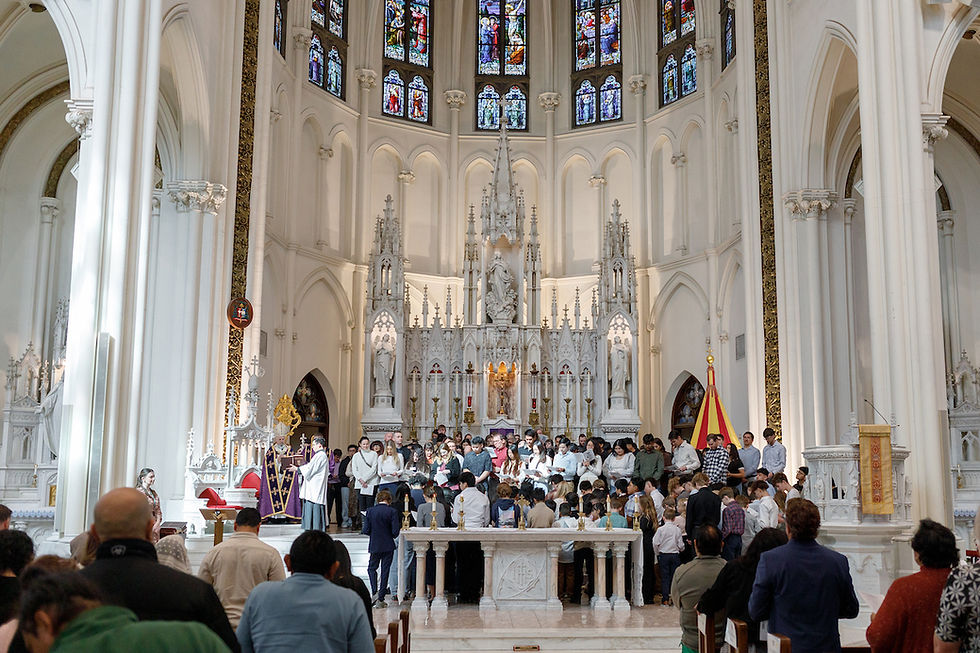 People gathered in a church near an ornate altar with stained glass windows. A priest addresses the congregation in a ceremonial setting.