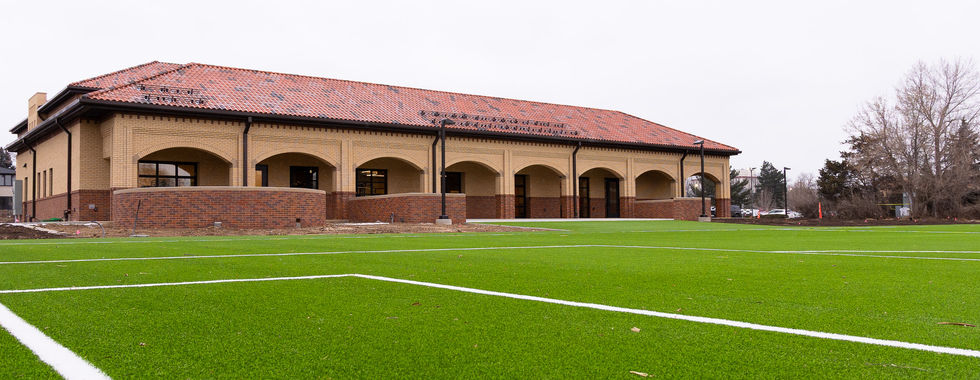 A building with arched windows and a red-tiled roof stands near a green field marked with white lines. Overcast sky and bare trees in the background.