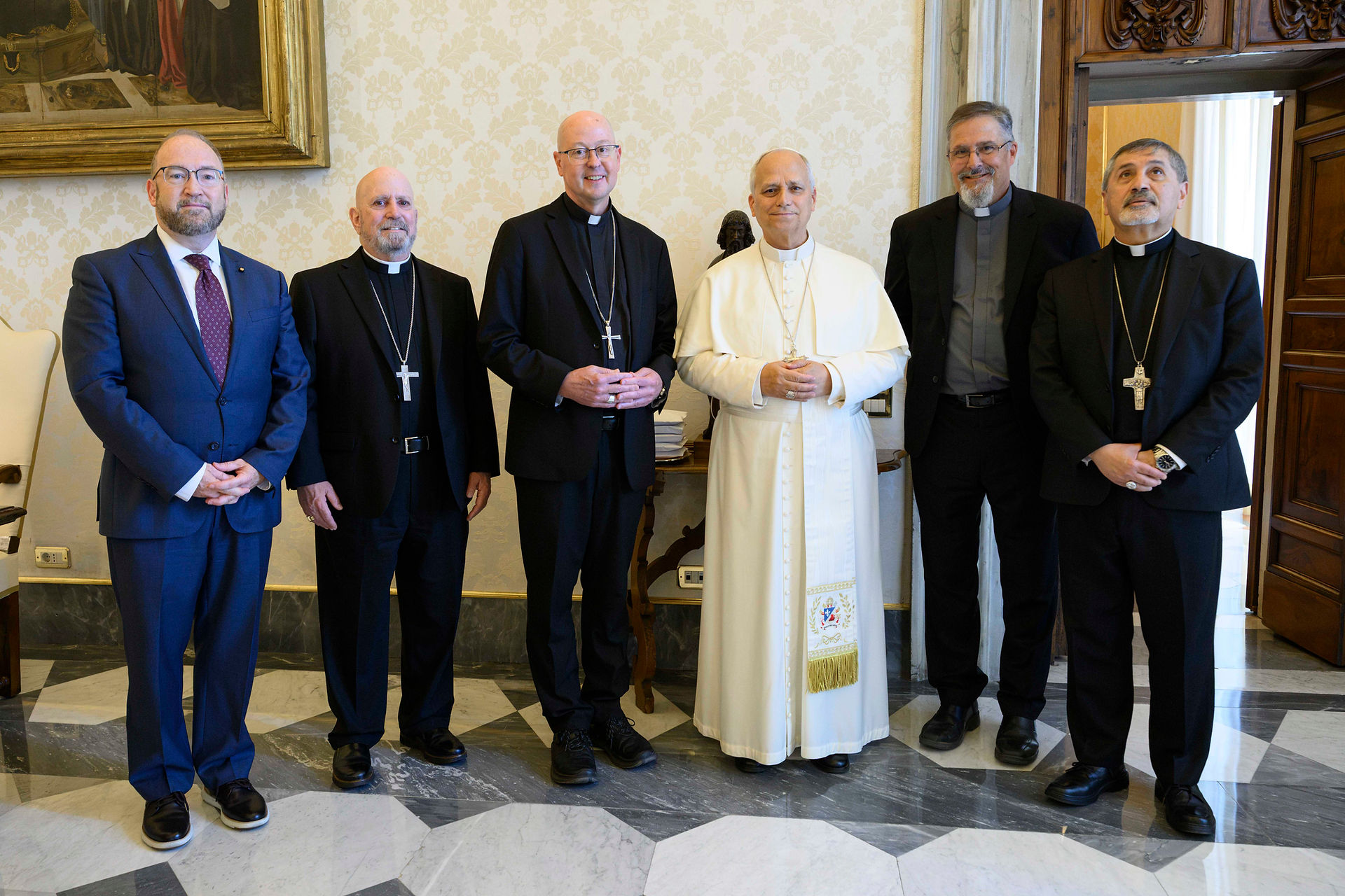 Six clergymen pose in a formal room, including the pope in white, all standing solemnly on a marble floor.