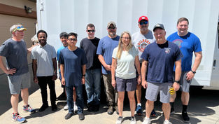 Group of 10 people pose smiling outdoors beside a white moving truck, with a sunny driveway and house in the background.
