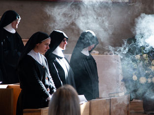 Nuns in black habits stand solemnly in a church amidst incense smoke, with sunlight highlighting their expressions, creating a serene atmosphere.
