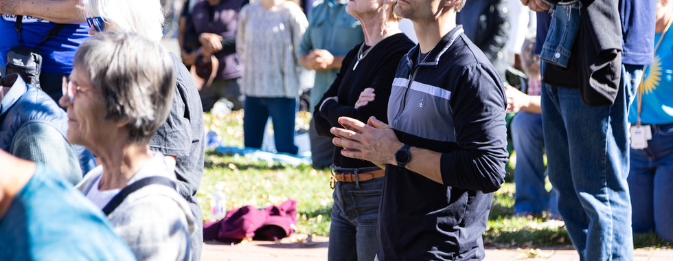 People are kneeling and standing in a park, praying. A man with glasses is in focus, wearing a black top. Sunlight illuminates the scene.