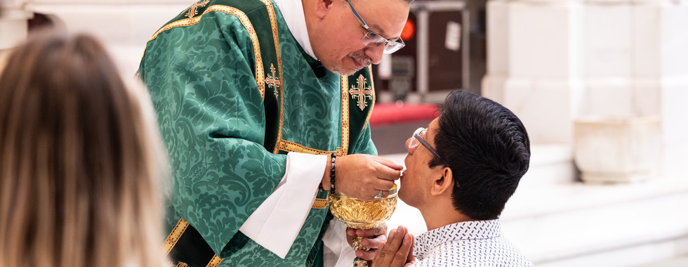 Deacon in green vestments gives communion with a gold chalice to a kneeling man in a bright church, solemn and reverent.