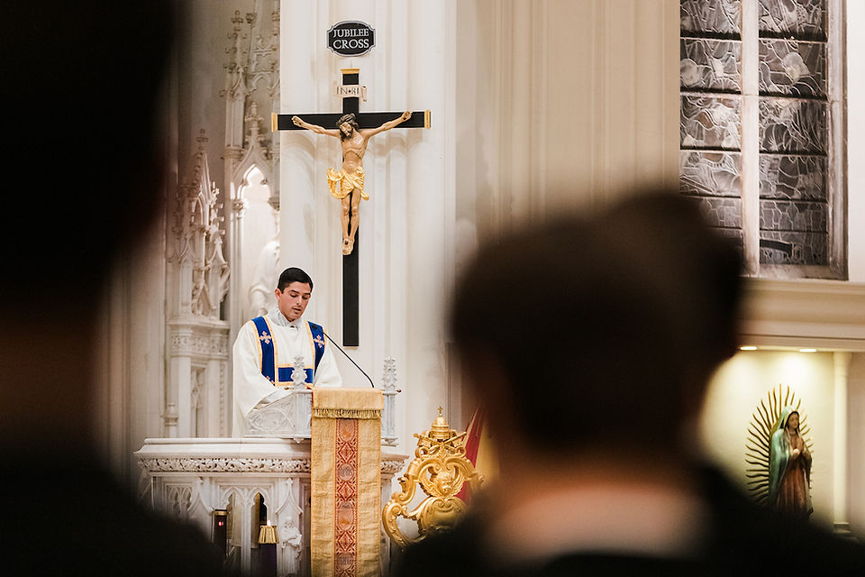 Deacon in blue and white robes at pulpit, reading in ornate church with crucifix and "Jubilee Cross" sign, solemn atmosphere.