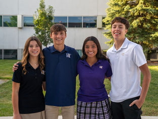 Four smiling students in colored polo shirts and plaid skirt, standing outdoors in front of a school building with trees in the background.