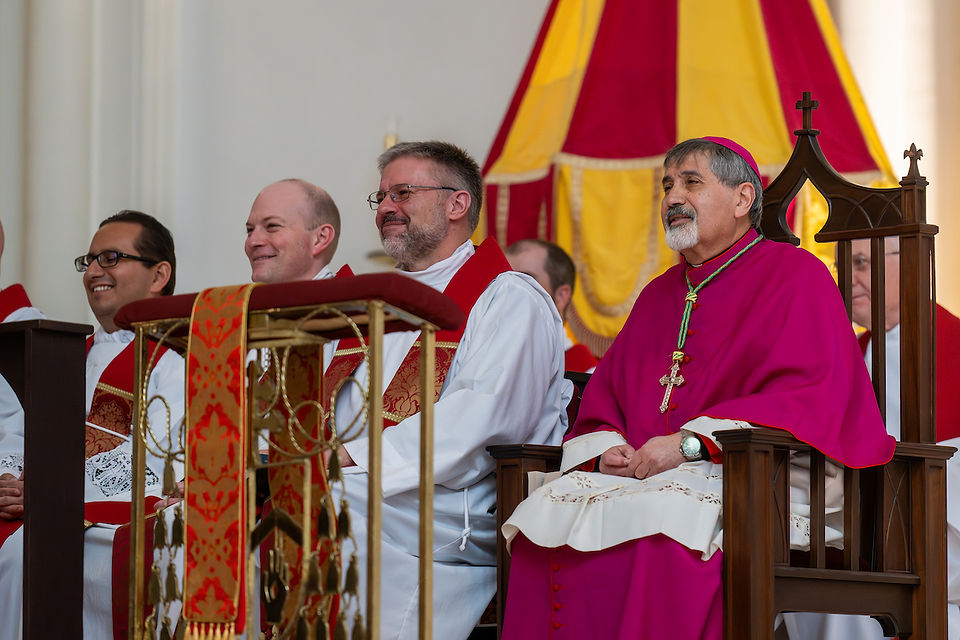 Clergy seated near the sanctuary, including a bishop in red vestments, listen attentively during the liturgy.