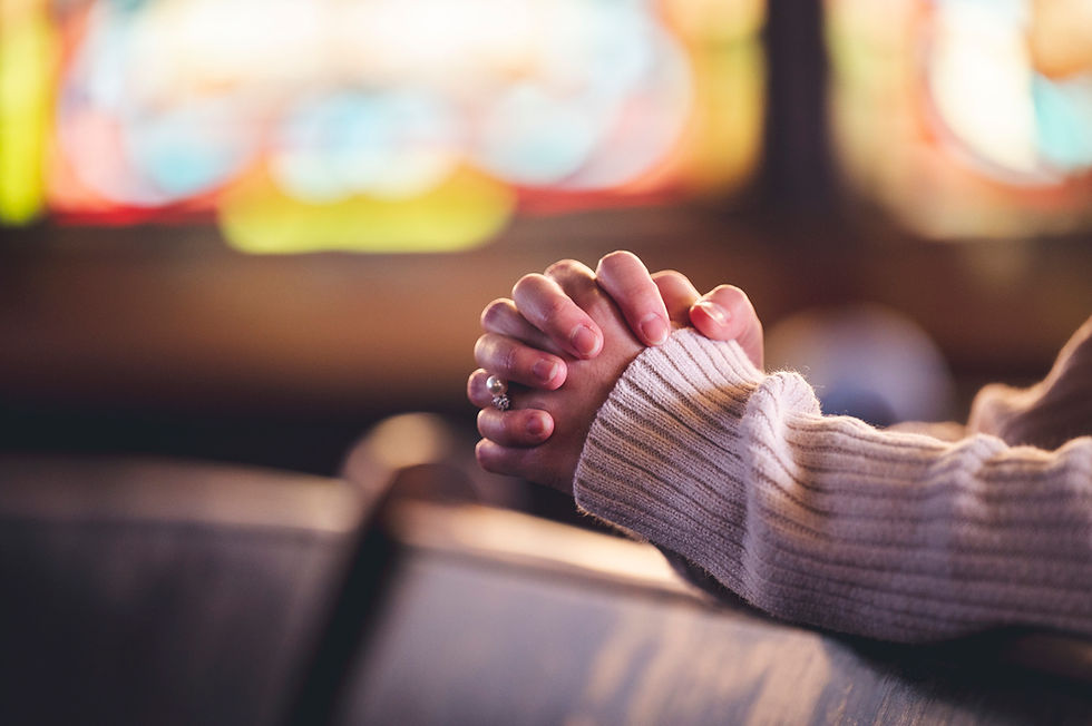 Hands clasped in prayer, resting on a wooden pew. A cozy sweater and blurred colorful stained glass background convey a peaceful mood.