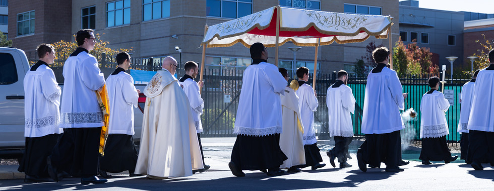 Priests in white robes participate in a religious procession outside a Planned Parenthood building. A golden canopy is carried above.