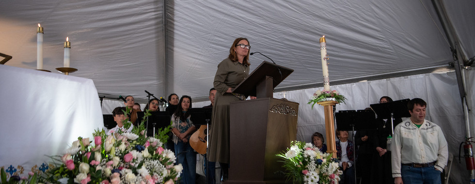 A woman speaks from a podium during the groundbreaking ceremony while musicians and attendees listen nearby.