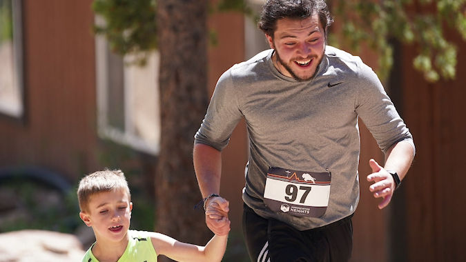 Man and child in running race hold hands, smiling. Child wears neon green shirt, man in gray. Race numbers 97 and 102 visible, outdoors.