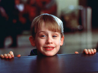 Young boy smiling over a table in a busy background, wearing a dark outfit. His hands grip the table's edge, creating a cheerful mood.