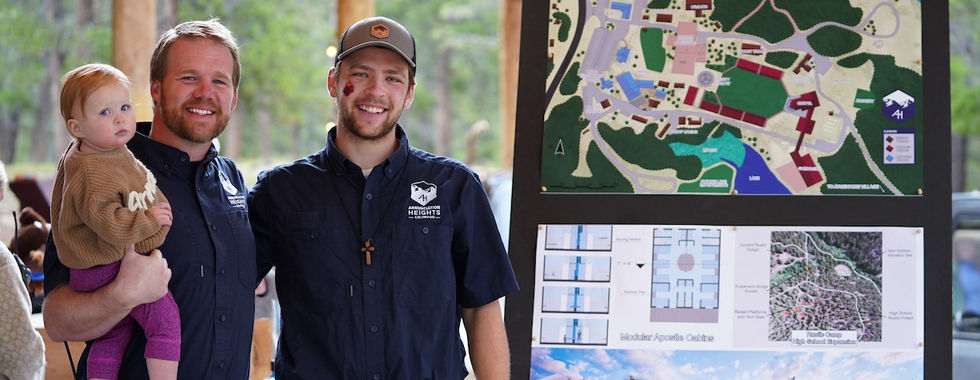 Two men smiling beside a large map display in a park pavilion. One holds a child in brown. They're wearing navy shirts with logos.