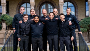 Nine men in clerical attire smile, posing together outdoors in front of an old building with arches. The mood is cheerful and relaxed.
