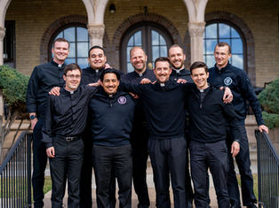 Nine men in clerical attire smile, posing together outdoors in front of an old building with arches. The mood is cheerful and relaxed.