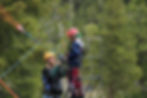 Man and child in helmets prepare for zipline on metal platform against lush forest background. Ropes and harnesses are visible.