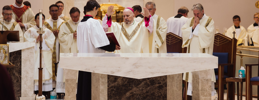 Clergy in ornate robes stand around an altar during a religious ceremony. One man raises hands in blessing. Stained glass in background.