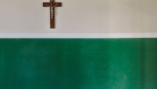 Crucifix on wall above green chalkboard with faint writing. Classroom setting with muted light and a contemplative, peaceful mood.