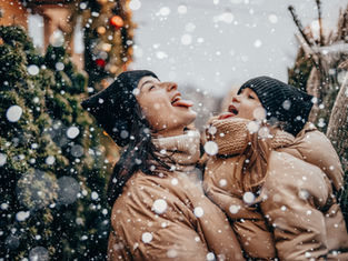 Two people in winter coats and hats catching snowflakes on their tongues. Snow falls against a background of evergreen trees. Joyful mood.
