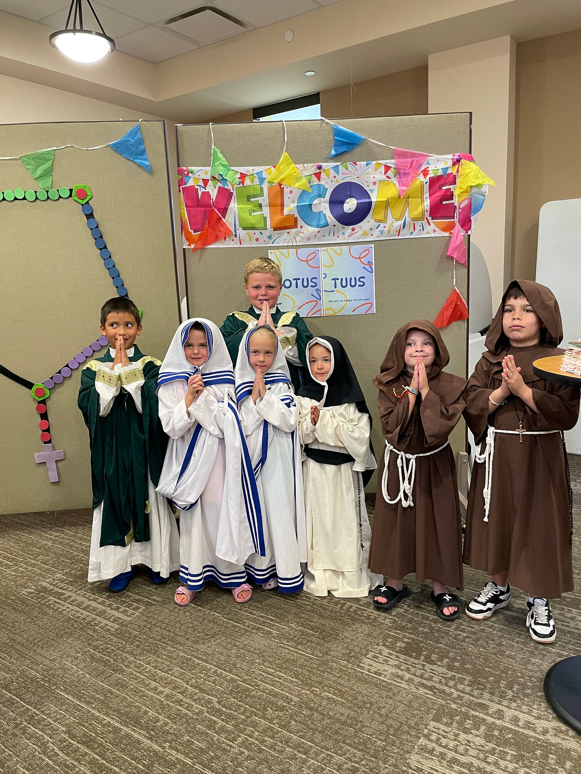 Children in religious costumes pose under a colorful "WELCOME" banner. Background features festive flags and a "TOTUS TUUS" sign.