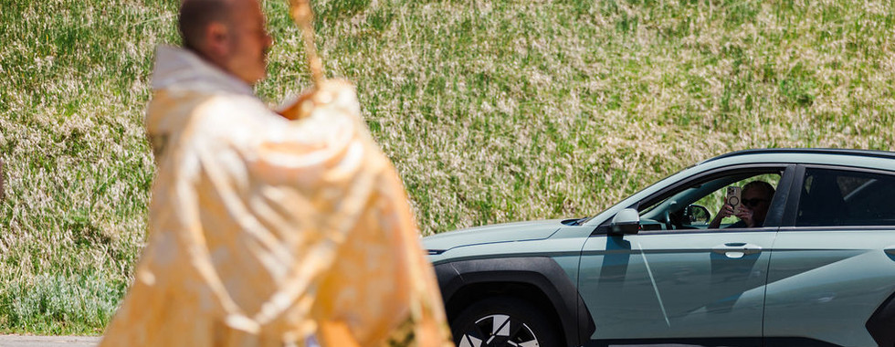 Blurred priest in gold robes stands by a green SUV as the driver takes a photo on a sunny roadside with grassy hill behind.