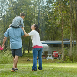 Family walking in a forest, with a boy on a man's shoulders and a girl holding his hand. A trailer and picnic table are in the background.