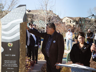 Man touches a stone memorial with an American flag design and the date May 7, 2019. People stand solemnly in a park setting.
