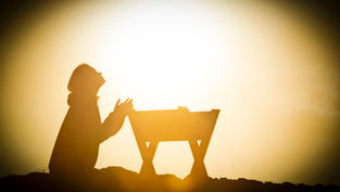 Silhouette of a person kneeling in prayer beside a manger at sunset, with golden light creating a peaceful, reverent mood.