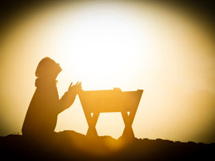 Silhouette of a person kneeling in prayer beside a manger at sunset, with golden light creating a peaceful, reverent mood.