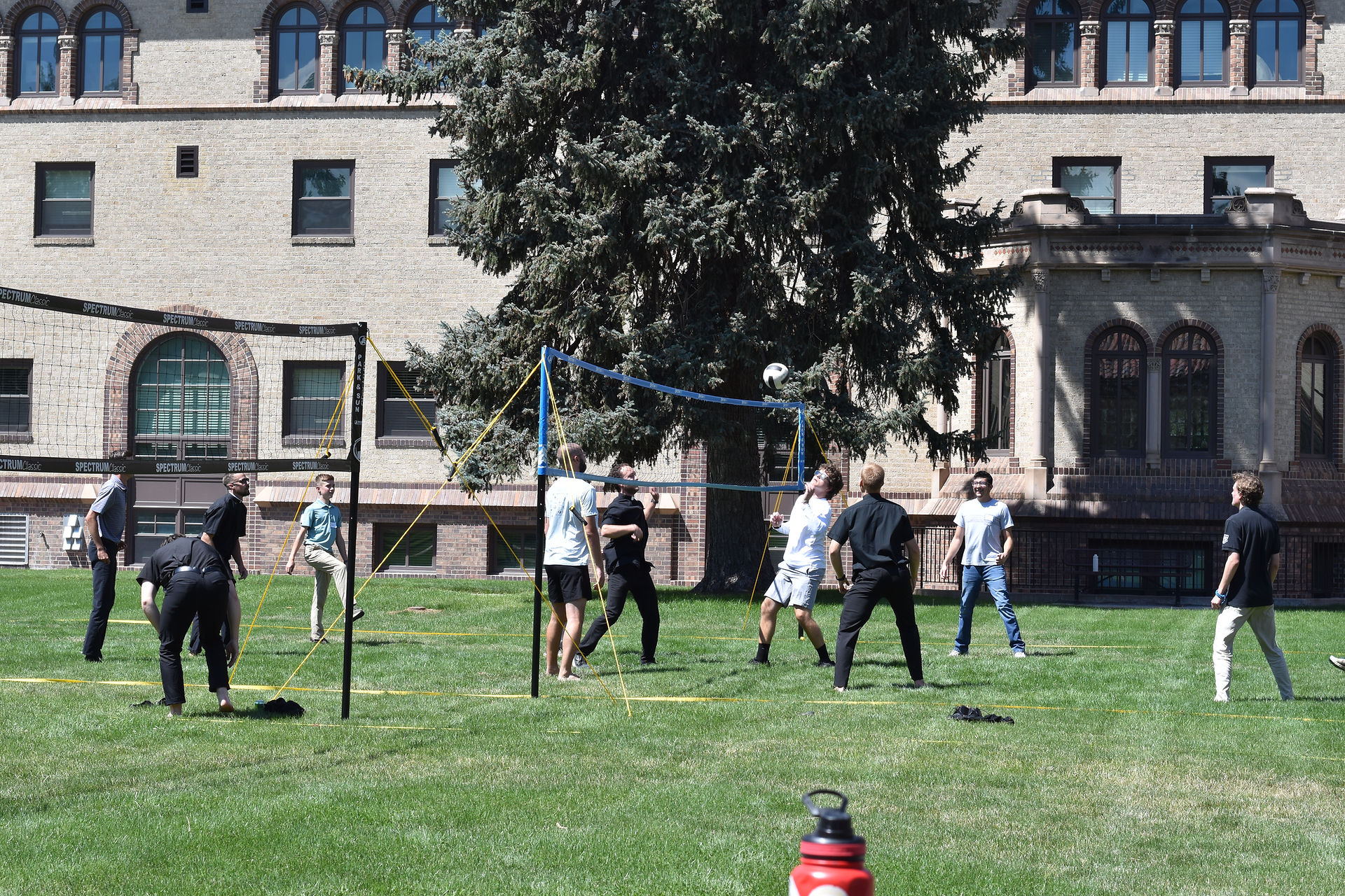 People playing volleyball on a grassy lawn by a brick building. A ball is mid-air, and a red water bottle is in the foreground.