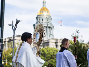 Procesión católica al aire libre: sacerdotes con una custodia dorada ante una cúpula dorada; cielo despejado y solemne.