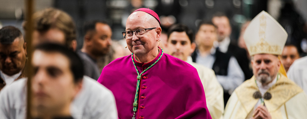 A bishop in a magenta robe smiles in a procession, surrounded by clergy in white and ornate attire. The background is blurred, giving a formal setting.
