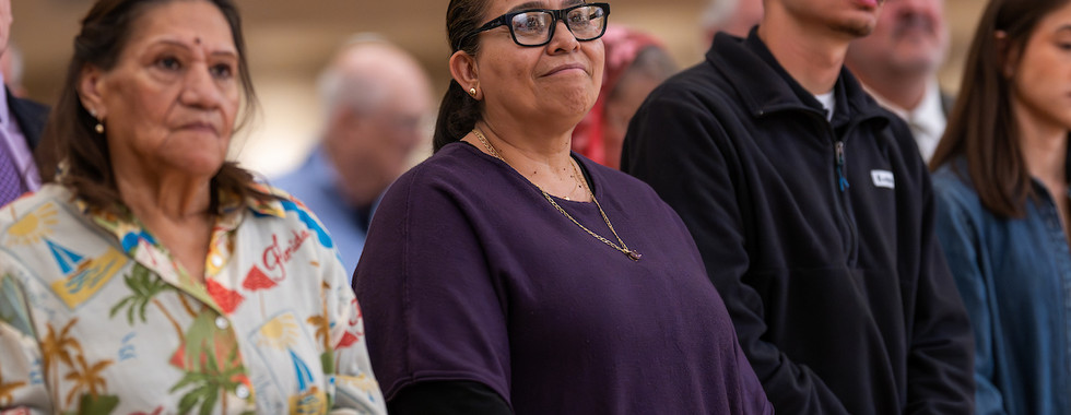 A group of people stands attentively indoors. A woman in purple is smiling, while others look focused. Background is softly blurred.