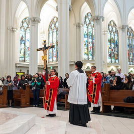 Clergy in red robes hold a large crucifix in a cathedral. Congregation stands in prayer. Stained glass windows in the background.