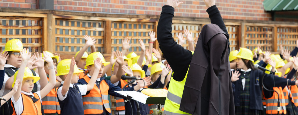 Children in hard hats and vests raise hands outside a brick building; adult leads with arms up, creating an energetic, organized scene.
