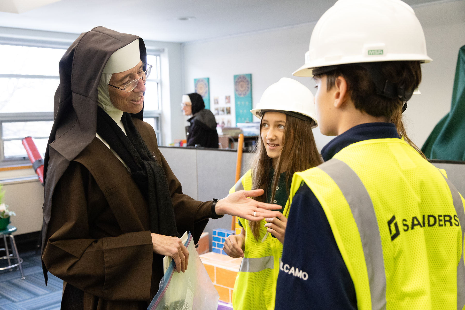 A nun in a brown habit smiles and gestures while talking to two children wearing white hard hats and yellow vests in a classroom setting.