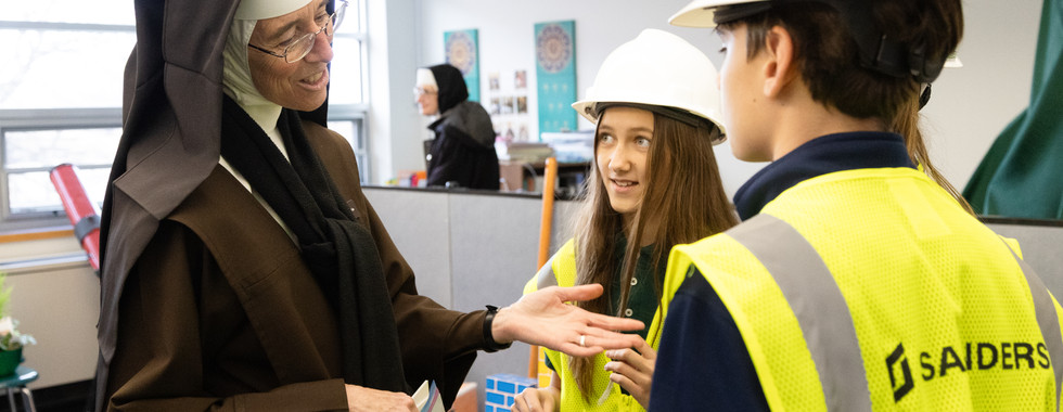 A nun in a brown habit smiles and gestures while talking to two children wearing white hard hats and yellow vests in a classroom setting.