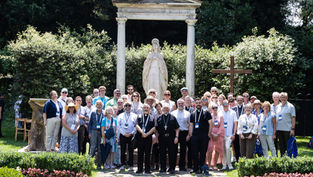 Group of tourists and priests pose before a white statue and cross in a sunlit garden.