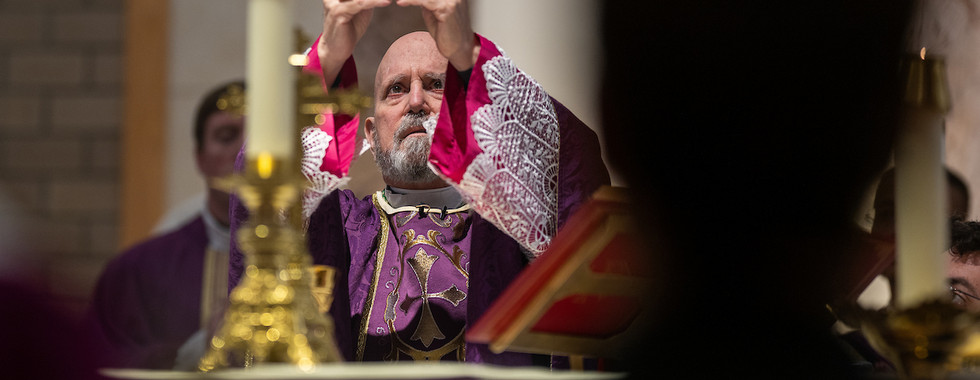 Bishop in purple vestments raises host during Mass. Candles and altar visible, solemn mood.