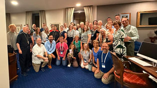 Large smiling group of adults, many with conference badges and clergy attire, posing together in a carpeted room with curtains and desk