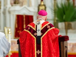 A clergyman in red and gold vestments, wearing a pink skullcap, stands at an altar. Another person in white robe beside him. Church setting.