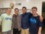 Four smiling friends pose indoors. One wears a light shirt with sunglasses print, another a plaid shirt, and two wear dark shirts with logos.