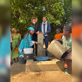 People and children gather at an outdoor hand pump, filling buckets and washing hands beneath leafy trees.