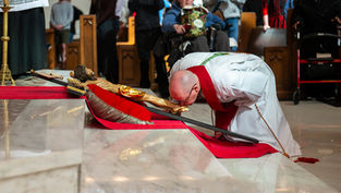 Obispo en vestiduras blancas y rojas besa un crucifijo en una iglesia, rodeado de fieles. Ambiente solemne, reflejos en el piso.