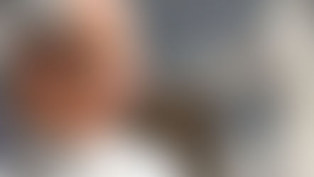 Smiling pope in white robes and skullcap, seen in profile against a blurred gray background, sunlit and peaceful