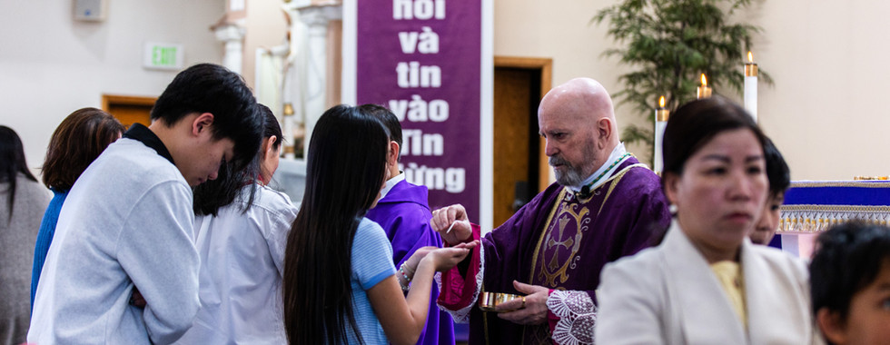 A bishop in purple robe gives communion in a church. People wait in line. A purple banner with text is visible in the background.